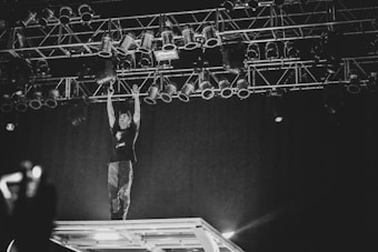 A performer stands on an elevated stage structure, reaching up towards a rigged lighting setup in a dimly lit environment. The atmosphere is dynamic and energetic, captured in black and white, with the focus on the interaction between the performer and the stage lights.