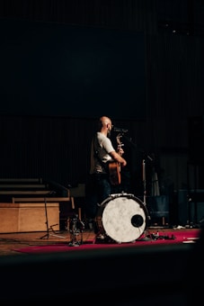 A musician is standing on a stage with a guitar, positioned in front of a microphone. There is a large drum in the foreground and various musical equipment scattered around. The lighting is low, giving a moody atmosphere to the scene.