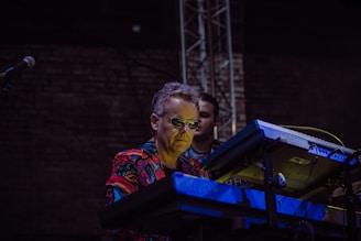 A musician wearing sunglasses and a colorful shirt plays an electronic keyboard on stage. The background is dark, with a brick wall and stage equipment visible.