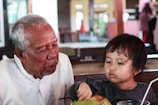 An elderly man and a young child are sitting together in a casual dining environment. The man appears focused on the child, who is reaching for a coconut drink with a straw. The scene captures a moment of interaction and connection.