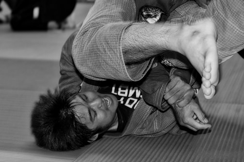 A person is grappling on a mat, possibly practicing a form of martial arts. The image is in black and white, showing the details of the gi uniform and physical exertion.