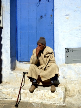 An elderly person is sitting on stone steps in front of a vibrant blue door. The individual is dressed in a beige, striped robe and a gray knit cap. A walking stick is leaning against the edge of the step. The wall surrounding the door is painted white, with some signs of weathering. There is a plaque with the number 204 near the bottom right of the image.