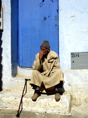 An elderly person is sitting on stone steps in front of a vibrant blue door. The individual is dressed in a beige, striped robe and a gray knit cap. A walking stick is leaning against the edge of the step. The wall surrounding the door is painted white, with some signs of weathering. There is a plaque with the number 204 near the bottom right of the image.