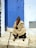 An elderly person is sitting on stone steps in front of a vibrant blue door. The individual is dressed in a beige, striped robe and a gray knit cap. A walking stick is leaning against the edge of the step. The wall surrounding the door is painted white, with some signs of weathering. There is a plaque with the number 204 near the bottom right of the image.