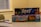 Two board game boxes are stacked on a table, with the titles 'Reef' and 'Tiny Towns' visible. A toy car and a magnifying glass rest on the table next to them. The background includes a window with blinds and part of a patterned chair.
