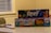 Two board game boxes are stacked on a table, with the titles 'Reef' and 'Tiny Towns' visible. A toy car and a magnifying glass rest on the table next to them. The background includes a window with blinds and part of a patterned chair.