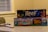 Two board game boxes are stacked on a table, with the titles 'Reef' and 'Tiny Towns' visible. A toy car and a magnifying glass rest on the table next to them. The background includes a window with blinds and part of a patterned chair.