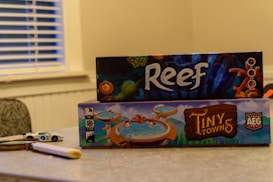 Two board game boxes are stacked on a table, with the titles 'Reef' and 'Tiny Towns' visible. A toy car and a magnifying glass rest on the table next to them. The background includes a window with blinds and part of a patterned chair.