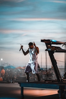 A musician is performing energetically on an outdoor stage, holding a microphone. The crowd in the background suggests a lively festival atmosphere. Musical equipment, like a keyboard, is visible on the stage. The sky is a mix of blue and gray hues, creating a dynamic backdrop.