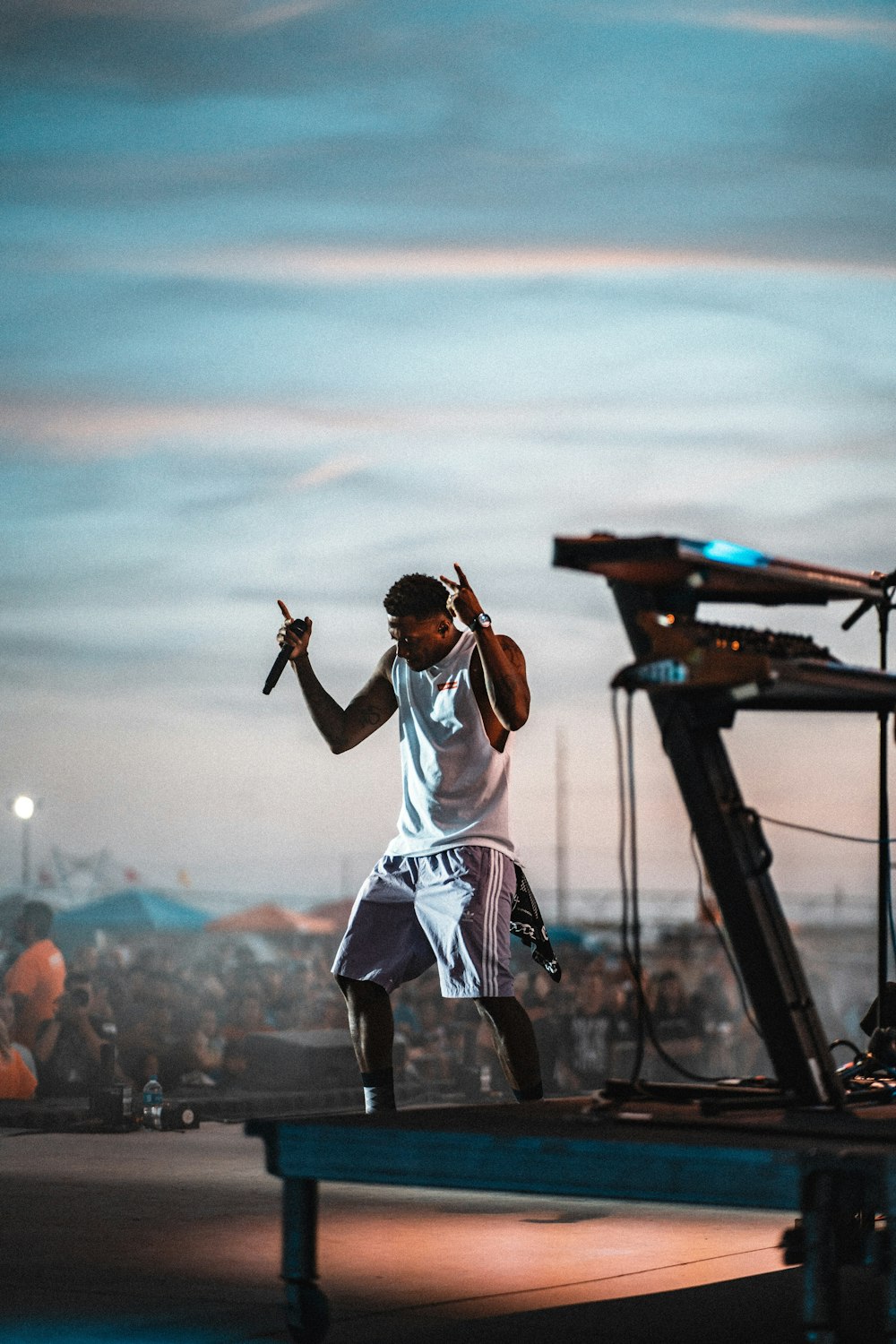 man wearing white tank top and gray shorts holding microphone while standing and performing on stage surrounded with people watching