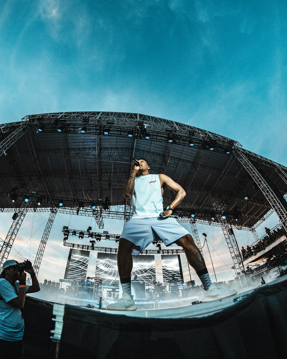 man wearing white tank top and shorts singing and performing on stage under blue and white skies during daytime