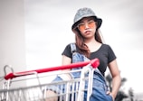 A woman wearing a patterned bucket hat, orange-tinted sunglasses, a black shirt, and denim overalls stands behind a shopping cart with a red handle. The background is cloudy, and the overall scene conveys a casual, urban vibe.