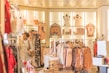 A well-lit clothing store featuring a variety of women's clothing items and accessories displayed on racks and shelves. The shelves include bags, mannequins with tops, and neatly hung dresses. A woman wearing a beige coat with red pants is browsing through the store.