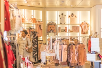 A well-lit clothing store featuring a variety of women's clothing items and accessories displayed on racks and shelves. The shelves include bags, mannequins with tops, and neatly hung dresses. A woman wearing a beige coat with red pants is browsing through the store.