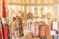 A well-lit clothing store featuring a variety of women's clothing items and accessories displayed on racks and shelves. The shelves include bags, mannequins with tops, and neatly hung dresses. A woman wearing a beige coat with red pants is browsing through the store.