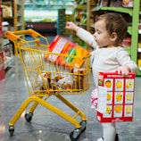 A toddler is inside a grocery store aisle, holding a colorful snack box in one hand, and placing another item into a small yellow shopping cart filled with various products. The shelves in the background are stocked with more grocery items.