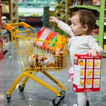 A toddler is inside a grocery store aisle, holding a colorful snack box in one hand, and placing another item into a small yellow shopping cart filled with various products. The shelves in the background are stocked with more grocery items.