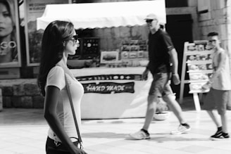 A black and white street scene captures a woman in profile with long hair and sunglasses walking past a market stall displaying handmade goods. Two people are seen walking in the opposite direction, with a backdrop of posters and a stone wall.