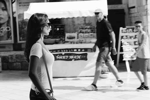 A black and white street scene captures a woman in profile with long hair and sunglasses walking past a market stall displaying handmade goods. Two people are seen walking in the opposite direction, with a backdrop of posters and a stone wall.