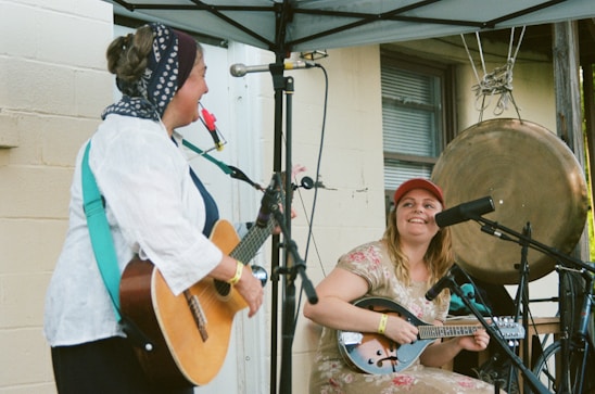 Two musicians are performing under a canopy. One is playing an acoustic guitar, wearing a headscarf, and has a red flower poking out of their mouth. The other is sitting with a mandolin, wearing a red cap, and smiling. A large gong is hanging in the background. Microphones are set up in front of both musicians.