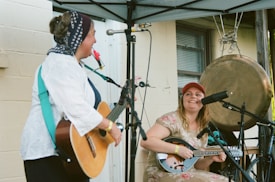 Two musicians are performing under a canopy. One is playing an acoustic guitar, wearing a headscarf, and has a red flower poking out of their mouth. The other is sitting with a mandolin, wearing a red cap, and smiling. A large gong is hanging in the background. Microphones are set up in front of both musicians.