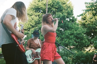 A band performs outdoors with a female singer wearing a red outfit holding a microphone. Two guitarists, one with long hair playing an electric guitar and another in a polka-dot dress playing a bass guitar, accompany her. Green trees fill the background, suggesting a park or garden setting.
