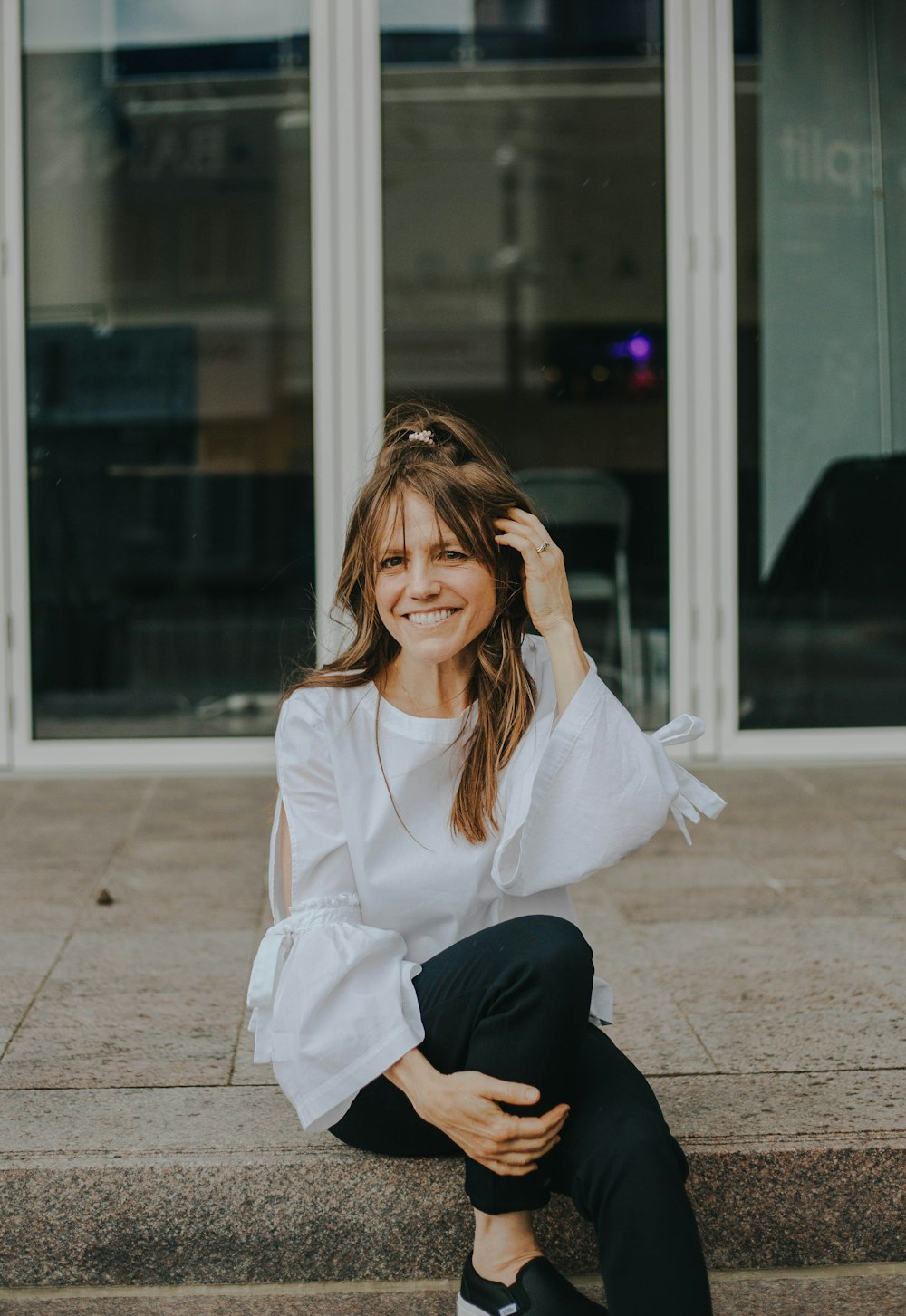 smiling woman touching her hair while sitting on concrete pathway near building