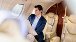 A person is seated comfortably inside a private jet, wearing a blue suit jacket and light-colored trousers. The interior features plush beige leather seats and a window through which soft light filters, creating a serene atmosphere. The cabin has elegant wood and gold-tone detailing.