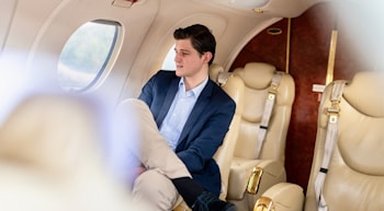 A person is seated comfortably inside a private jet, wearing a blue suit jacket and light-colored trousers. The interior features plush beige leather seats and a window through which soft light filters, creating a serene atmosphere. The cabin has elegant wood and gold-tone detailing.