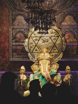 A large statue of Ganesha, adorned with vibrant clothes and gold accessories, stands in front of an ornately decorated backdrop. The setting is illuminated by a grand chandelier, and the background features Hindu symbols and intricate patterns. Silhouettes of people are visible in the foreground.