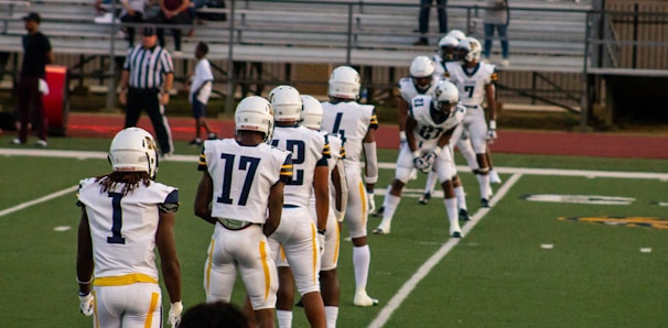 A group of American football players in white and navy uniforms are lined up on a green field, preparing for a play. Their helmets are white, and they are positioned near the sideline with a referee and spectators visible in the background.