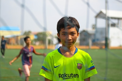 A young boy is wearing a bright yellow soccer jersey with a Barcelona logo, standing on a green soccer field. In the background, there are other children blurred in motion, indicating they are playing. The setting appears to be outdoors with clear skies and distant buildings.