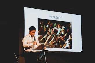 A man in a white shirt stands at a lectern holding a microphone. Behind him is a large screen displaying an image of a crowd raising their hands with the word 'WORSHIP' above it.