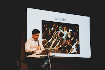 A man in a white shirt stands at a lectern holding a microphone. Behind him is a large screen displaying an image of a crowd raising their hands with the word 'WORSHIP' above it.