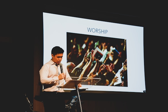 A man in a white shirt stands at a lectern holding a microphone. Behind him is a large screen displaying an image of a crowd raising their hands with the word 'WORSHIP' above it.