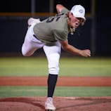 A baseball player is in the middle of a pitch on a baseball field. The player is wearing a green shirt, white pants, a white cap, and a gold chain. The background shows green grass and the outlines of the baseball field.