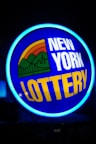 A bright, circular neon sign with a blue background, featuring the words 'New York Lottery' in bold white and yellow letters. The sign includes a colorful illustration of a landscape with green mountains, an orange-red sunset, and a bridge, all encapsulated within the circle.