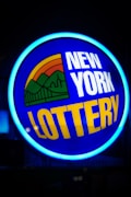 A bright, circular neon sign with a blue background, featuring the words 'New York Lottery' in bold white and yellow letters. The sign includes a colorful illustration of a landscape with green mountains, an orange-red sunset, and a bridge, all encapsulated within the circle.