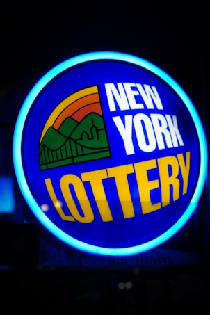 A bright, circular neon sign with a blue background, featuring the words 'New York Lottery' in bold white and yellow letters. The sign includes a colorful illustration of a landscape with green mountains, an orange-red sunset, and a bridge, all encapsulated within the circle.