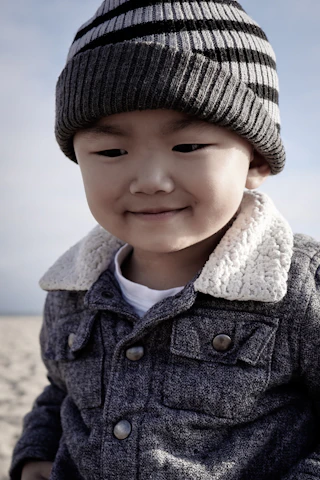 A young child wearing a striped knit hat and a textured jacket with a shearling collar is outdoors. The child has a gentle smile and appears to be at the beach, with sand visible in the background under a clear sky.
