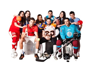A group of eight young adults, each dressed in different sports uniforms, are posed together smiling. They hold various sports equipment including a soccer ball, volleyball, basketball, football, tennis racket, and table tennis paddle. The clothing includes jerseys and gear for soccer, hockey, football, and other sports. The background is plain white, emphasizing the colorful attire of the group.