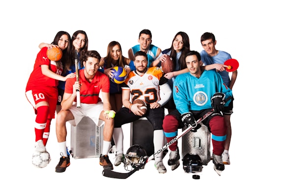 A group of eight young adults, each dressed in different sports uniforms, are posed together smiling. They hold various sports equipment including a soccer ball, volleyball, basketball, football, tennis racket, and table tennis paddle. The clothing includes jerseys and gear for soccer, hockey, football, and other sports. The background is plain white, emphasizing the colorful attire of the group.