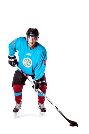 A hockey player is poised in a ready stance, wearing a blue jersey with a crest on the chest, black shorts with red socks, black gloves, and a black helmet with a clear visor. He is holding a hockey stick on the white, smooth ice surface.