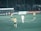 A group of soccer players is on a field, with several players wearing yellow and green uniforms and one in a white uniform. The field is surrounded by advertisements with a crowd behind the fence.
