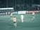 A group of soccer players is on a field, with several players wearing yellow and green uniforms and one in a white uniform. The field is surrounded by advertisements with a crowd behind the fence.