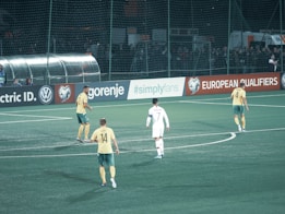 A group of soccer players is on a field, with several players wearing yellow and green uniforms and one in a white uniform. The field is surrounded by advertisements with a crowd behind the fence.