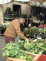 An older man wearing a brown sweater and a black hat is shopping at an outdoor market. He is examining a variety of fresh greens and vegetables, including leafy greens and eggplants. The market stalls are filled with abundant produce, and other shoppers are visible in the background.