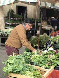 An older man wearing a brown sweater and a black hat is shopping at an outdoor market. He is examining a variety of fresh greens and vegetables, including leafy greens and eggplants. The market stalls are filled with abundant produce, and other shoppers are visible in the background.