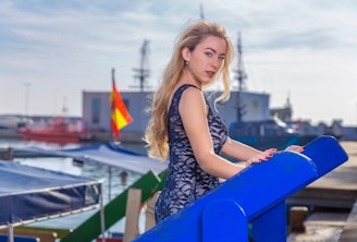 A woman with long blonde hair and a navy lace dress stands at the edge of a blue railing near a harbor. The background displays a shipyard with buildings, cranes, and a blurred Spanish flag. The sky is mostly clear with soft sunlight illuminating the scene.