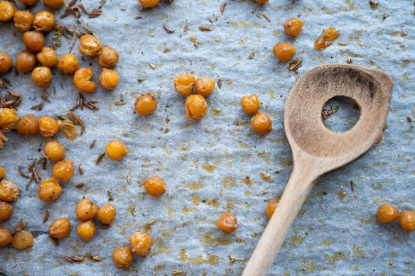 A wooden spoon rests on baking paper scattered with crunchy roasted chickpeas and seasoning flakes, creating a rustic and homemade vibe.